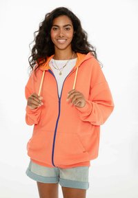 Korálová mikina s na zips a oranžovým vnútrom, modrým zipsom. Má voľný strih, veľké predné vrecká a šnúrky. Casual šortky.