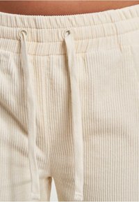 Cremfarbene Cordhose mit gerippter Textur, ausgestattet mit einem Kordelzugbund und Metallösen. Der Stoff zeigt dezente vertikale Linien.