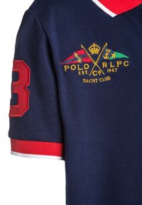 Marinflöjt med röd krage och ärmslut. Har en broderad logotyp med korsade flaggor och guldtext. Röd nummer "3" på ärmen.