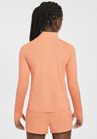 Langärmliges Sportoberteil und Shorts aus leichtem orangefarbenem Stoff, mit hohem Kragen, glatter Textur und taillierter Passform.