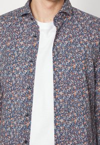 Man in een ongebotonde blauwe shirt met kleine oranje en witte bloemenprint over een eenvoudige witte T-shirt met ronde hals.