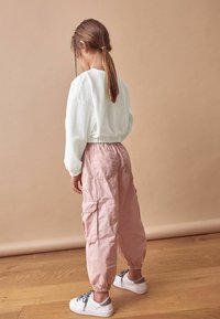 Jasnozielony, fakturowany sweter, różowe spodnie cargo z elastycznymi mankietami i białe sneakersy ze srebrnymi akcentami, widoczne z tyłu na drewnianej podłodze.