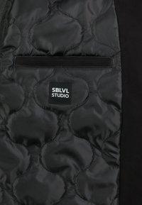 De binnenkant van de zwarte gewatteerde jas heeft een soepele stof met een cirkelpatroon. Bevat een zak en een aangehechte "SBLVL STUDIO" label.