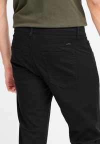 Pantalons en denim noir avec une taille mi-haute, poches arrières plates avec un subtil détail de couture en chevron, et une texture lisse et mate.