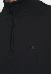 Sudadera negra con cremallera y cuello alto, hecha de tejido acanalado. Presenta un sutil logo "HUGO" en un color a juego en el pecho.