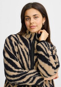 Veste en polaire zippée couleur beige avec des rayures noires de tigre ; elle présente un col haut, un tissu texturé et des fermetures éclair et poignets noirs contrastants.