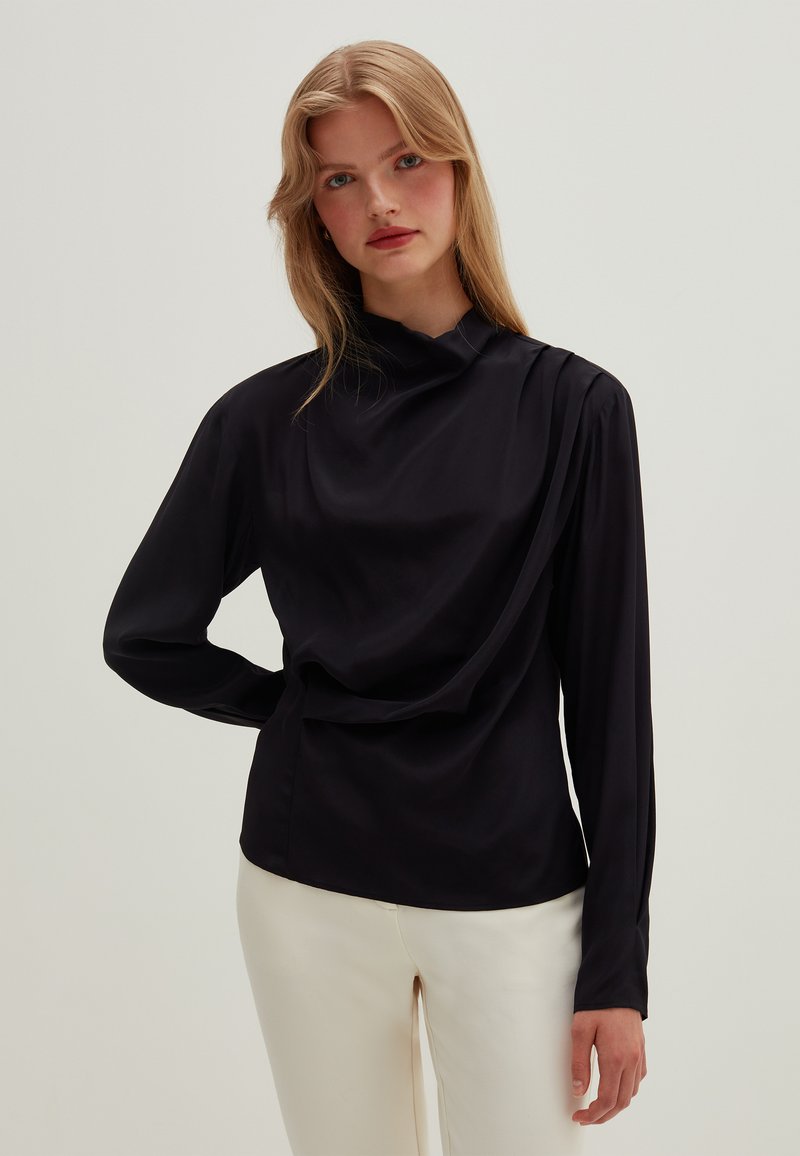 Zwarte blouse met lange mouwen, hoge halslijn en gedrapeerd ontwerp, gemaakt van een gladde stof. Gecombineerd met lichtgekleurde broeken.