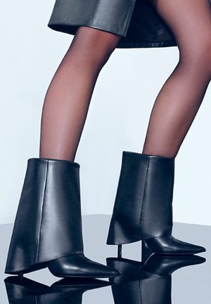 Botines de cuero negro hasta el tobillo con punta afilada, tacón alto y ancho, y un puño distintivo, reposando sobre una superficie reflectante.