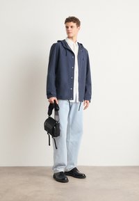 Navy hooded jacket with front buttons, paired with a white shirt, light blue jeans, and black shoes, holding a small black bag with a strap.