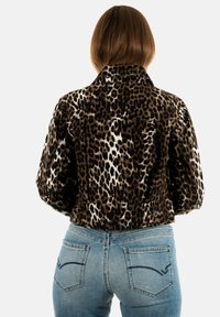 Donna con capelli castani di lunghezza media che indossa una giacca leopardata corta e jeans in denim azzurro chiaro, vista da dietro.