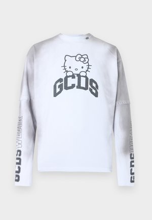 Mężczyzna stojący w szarych jeansach o szerokich nogawkach, białej koszulce z długim rękawem z napisem Hello Kitty i GCDS oraz czarnych butach na jednolitym jasnym tle.