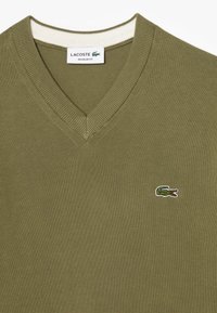 Suéter verde oliva de cuello en V con cuello acanalado, que presenta un pequeño logo de cocodrilo bordado en verde y rojo en el lado izquierdo del pecho. Corte regular.