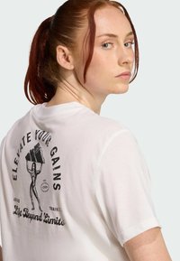 T-shirt en coton blanc avec un graphique vintage au dos, portant le texte "AMÉLIOREZ VOS GAINS," et un design représentant une femme soulevant des poids.