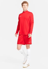 Rode sportieve trainingspak van gladde stof, met een top met halve rits en bijpassende shorts, gecombineerd met witte sokken en voetbalschoenen.