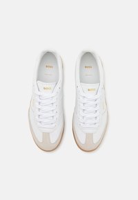 Baskets en cuir blanc avec des accents en daim beige, dotées de lacets blancs, d'une semelle texturée et d'un logo doré sur la languette et le côté.