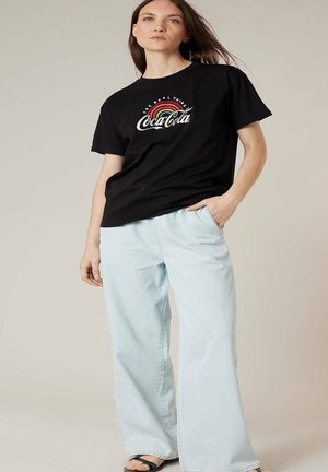 Femme portant un T-shirt Coca-Cola noir avec un logo arc-en-ciel, un jean large bleu clair et des sandales noires, debout avec une main dans la poche.