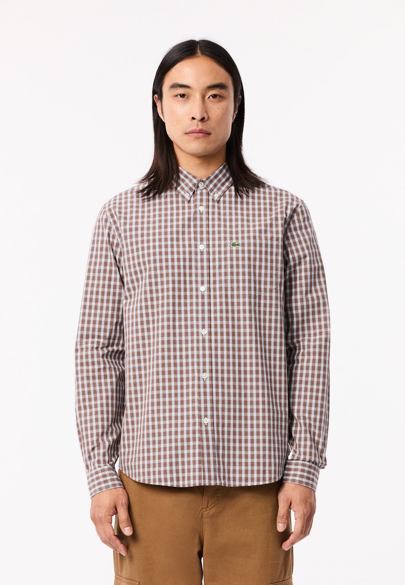 Chemise à carreaux brune et blanche à boutons avec un col, des manches longues, une poche poitrine et un petit logo vert sur le côté gauche de la poitrine.