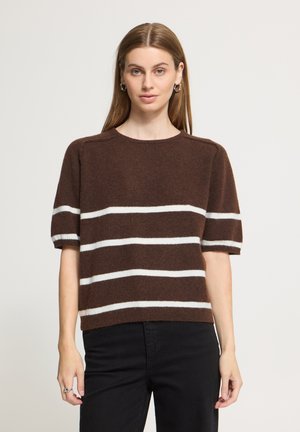 Femme debout, de face, portant un pull marron à manches courtes avec des rayures horizontales blanches et un pantalon noir sur fond uni.