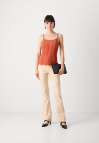 Top en tricot orange avec un ourlet festonné et des bretelles réglables, associé à un pantalon beige évasé et des chaussures à talons noires, tenant une pochette noire.