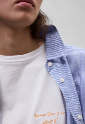 Nahaufnahme einer Person, die ein weißes T-Shirt mit orangefarbenem Text unter einem blau-weiß gestreiften Hemd trägt.