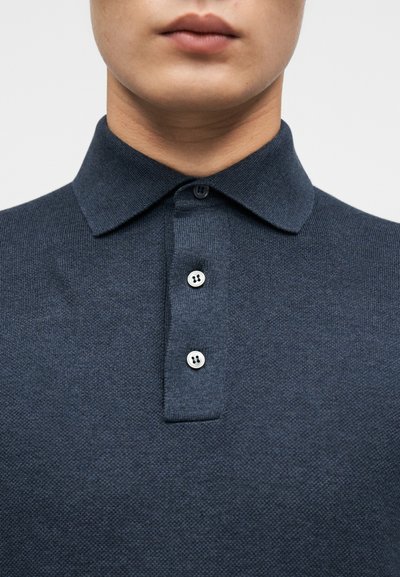Σκούρο μπλε πόλο πουκάμισο από πλεκτό ύφασμα. Διαθέτει κλασικό γιακά και τριπλή πατιλέτα με στρογγυλά, αντίθετου χρώματος κουμπιά.