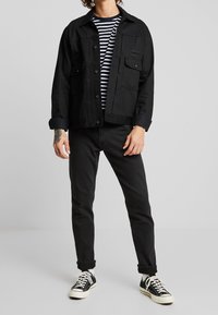 Schwarze Jeansjacke mit Knopfleiste, zwei Brusttaschen, getragen über einem schwarz-weiß gestreiften T-Shirt, kombiniert mit schwarzer Jeans und Sneakern.