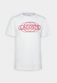 Bílý bavlněný tričko s červeným logem "LACOSTE" na přední straně, krátké rukávy a kulatý výstřih. Hladká textura.