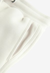Detail blízkého záběru na bílý elastický pas a boční kapsu na měkkých bavlněných kalhotách proti světlému pozadí.