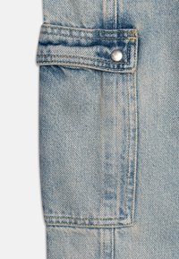 Jasnoniebieska tkanina dżinsowa, z założoną kieszenią z zapięciem na napę. Widoczne zszycia i teksturowane wykończenie na całej powierzchni.