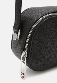 Primo piano di un angolo di una borsa in pelle nera con trama, con cerniera argentata e un tirante della cerniera a forma di spilla da balia argentata attaccato alla tracolla.