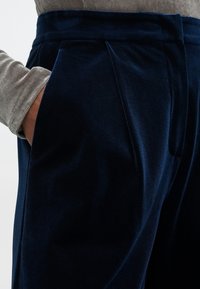 Pantalons en velours bleu marine avec un design plissé, des poches latérales et une texture lisse. Les détails comprennent une coupe ajustée et un doux éclat.