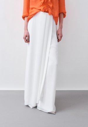 Osoba ubrana w jasnopomarańczową bluzkę z marszczonymi detalami i białe, szerokie spodnie o luźnym kroju, stojąca na szarej podłodze na tle białej ściany.