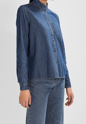 Person iført en langærmet blå denimskjorte med knapper og matchende blå denimjeans, stående mod en ensfarvet baggrund.