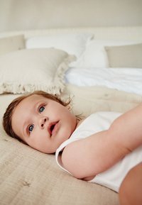 Un bebeluș îmbrăcat într-un body alb fără mâneci este întins pe o suprafață bej texturată, cu perne și lenjerie de pat deschise la culoare în fundal.