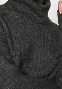 Mörkgrå ribbad stickad polotröja som bärs på överkroppen med en hand som rör vid nacken.