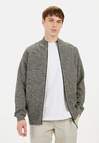 Graue, strukturierte Bomberjacke mit hohem Kragen und Reißverschluss, kombiniert mit einem weißen T-Shirt und hellen Hosen.