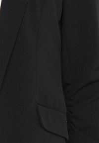 Czarna tkanina w zbliżeniu, pokazująca dopasowaną marynarkę z klapką i marszczonym detalem rękawa na prawym ramieniu.