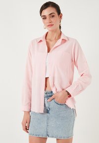 Light pink button-up shirt with a pocket, made of a textured fabric. Worn over a white crop top, paired with a blue denim skirt.