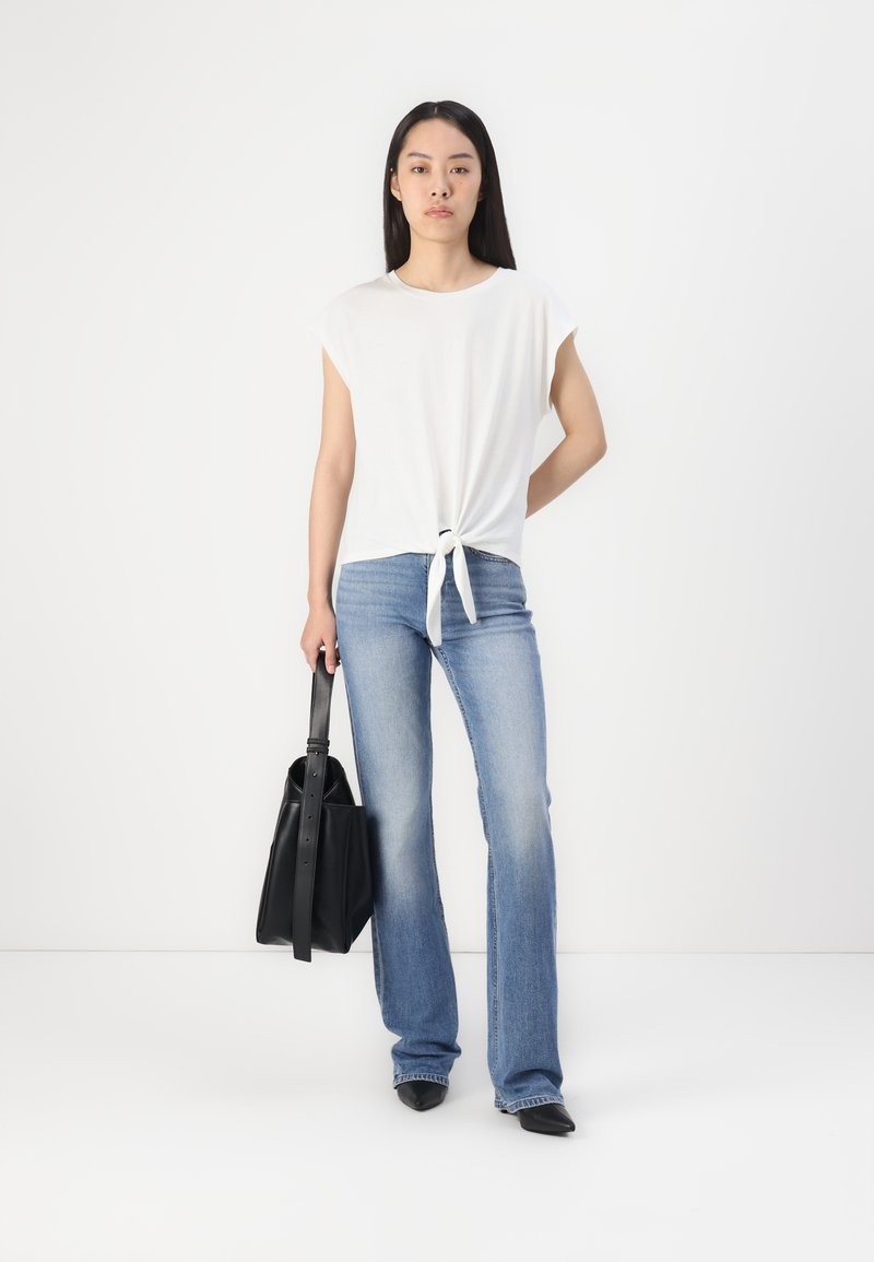 Weißes Kurzarmoberteil mit Knotendetail auf der Vorderseite, kombiniert mit hellblauen Schlagjeans. Das Model hält eine schwarze strukturierte Tasche.