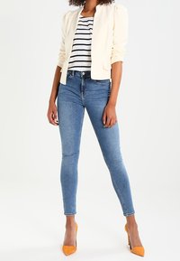 Blazer chiaro con maniche a sbuffo sopra una maglia a righe, abbinato a jeans skinny blu scuro e scarpe col tacco gialle.