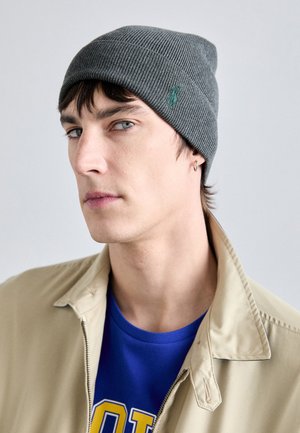 Gorro de punto acanalado gris con un sutil logo verde, llevado con una chaqueta beige sobre una camiseta azul. Textura suave con un ajuste ceñido.