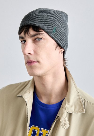 Gorro de punto acanalado gris con un sutil logo verde, llevado con una chaqueta beige sobre una camiseta azul. Textura suave con un ajuste ceñido.