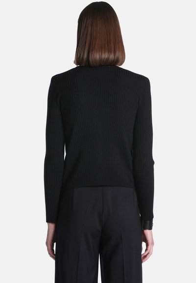 Femme aux cheveux bruns raides portant un haut noir côtelé à manches longues et un pantalon noir, vue de dos sur un fond uni.