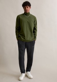 Maslinasto zelena majica dugih rukava s visokim ovratnikom u kombinaciji sa crnim trenirama i bijelim tenisicama. Ima jednostavan dizajn s suptilnim detaljima brendiranja.