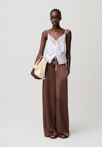 Mujer con top blanco sin mangas con encaje, pantalones marrones sueltos, sosteniendo un bolso beige, de pie frente a un fondo liso y claro.