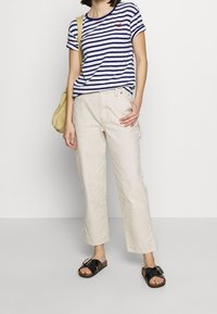 Camiseta de rayas en azul marino y blanco, pantalones beige de corte holgado y sandalias negras con hebilla grande. El modelo lleva un bolso de hombro amarillo.