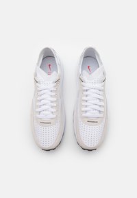 Bílé atletické boty s semišovými akcenty, perforovan�ým designem a bílými tkaničkami. Podešev má texturovanou černou spodní část. Viditelné červené logo.