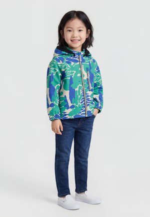 Enfant souriant portant une veste à capuche verte et bleue à motifs, un jean bleu foncé, et des chaussures blanches sans lacets, debout avec une main dans la poche.