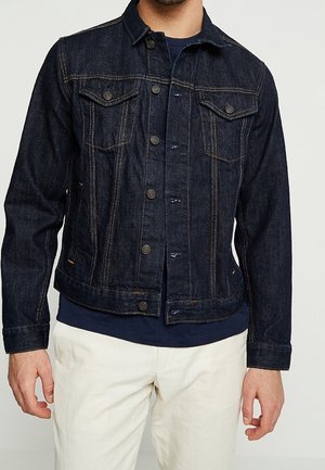 Mann trägt eine dunkelblaue Jeansjacke mit Knopfleiste, ein marineblaues Hemd darunter und cremefarbene Hosen, steht vor einem einfarbigen Hintergrund.