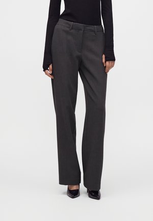 Femme portant un pantalon droit gris foncé, des talons noirs à bout pointu et un haut noir ajusté à manches longues avec des trous pour les pouces.